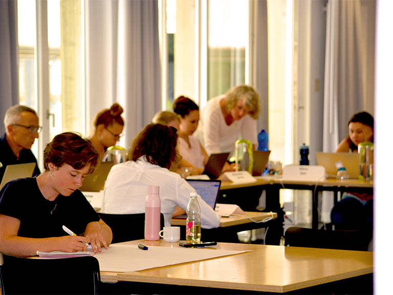 Zu sehen ist ein heller Kursraum mit Teilnehmenden die an den Tischen sitzen und arbeiten.