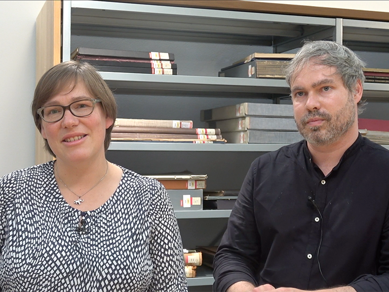 Zu sehen sind 2 Dozierende vor einem Bücherregal in einem Interviewshot.