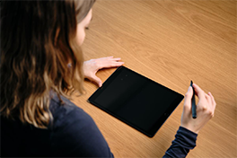 Studierende mit Headphone hält ein Tablet in den Händen und ist am Kommunizieren mit anderer Person. Auf dem Tisch steht eine durchsichtige Wasserflasche, daneben eine weisse Tasse.
